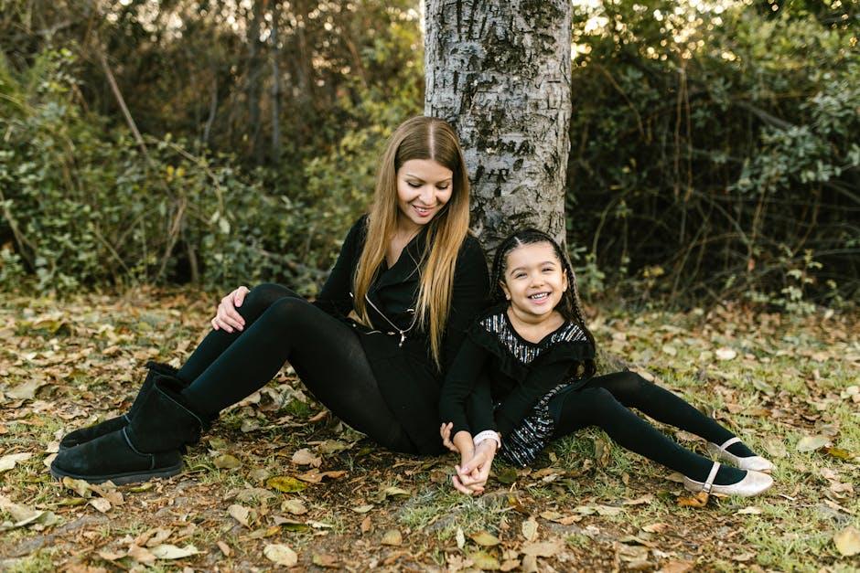 A mother and daughter sitting and smiling by a tree in a leafy park.