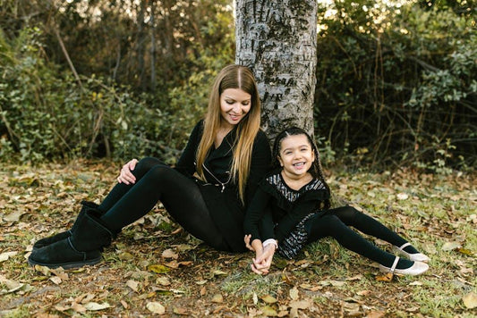 A mother and daughter sitting and smiling by a tree in a leafy park.