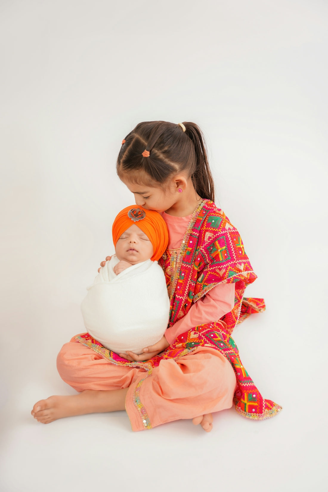 Older sister kisses newborn baby brother wearing an orange turban.