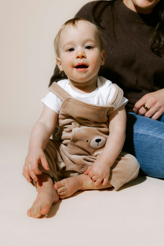 A baby wearing a bear-themed jumpsuit sits on the floor.