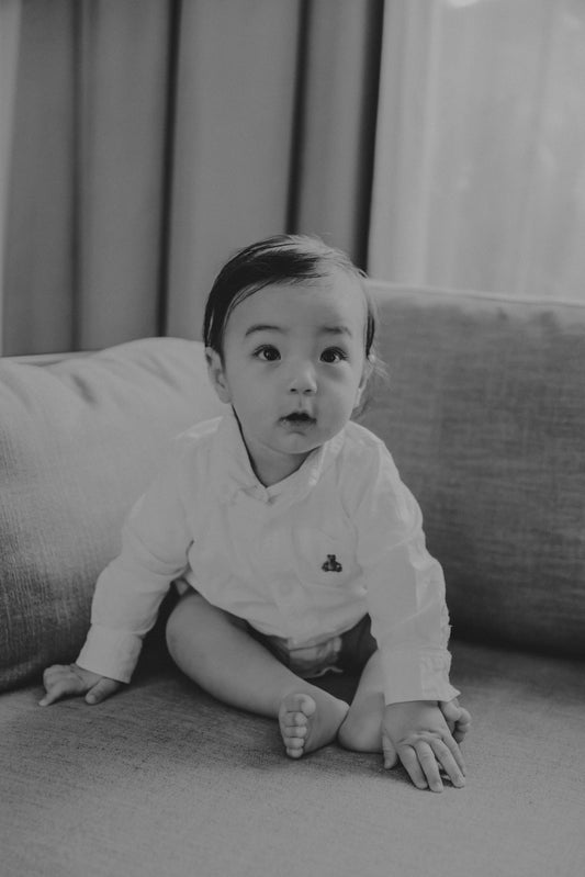A baby in a white shirt sits on a couch.