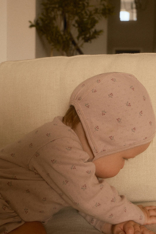 A baby in a pink bonnet leaning over a sofa.