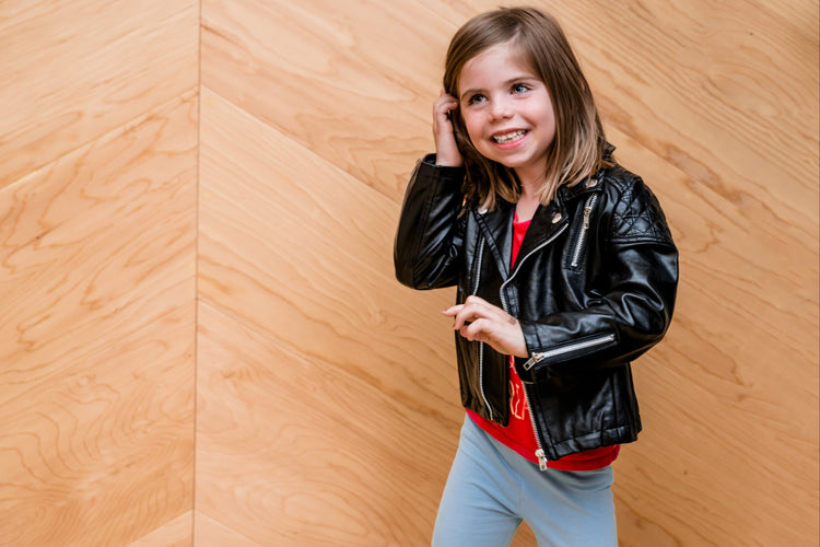 Smiling young girl in black biker jacket and light blue pants standing against wooden wall background