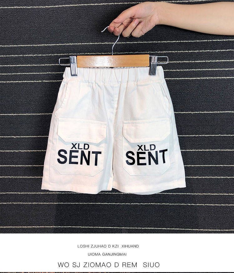 White kids shorts with back pockets labeled XLD SENT, held on wooden hanger over striped black background