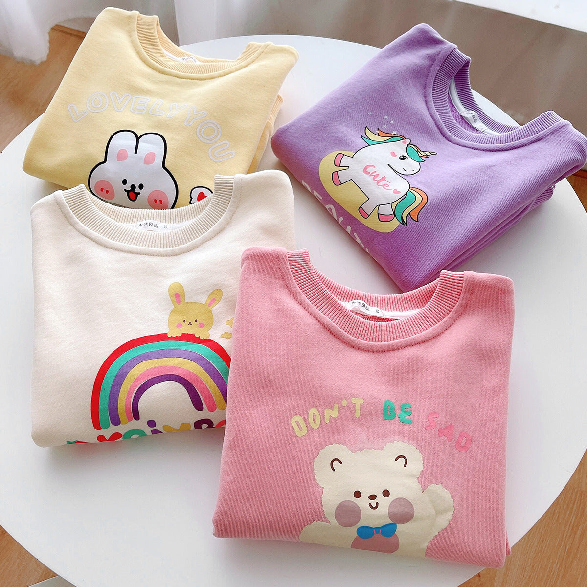 Four folded colorful children's sweatshirts with cute animal and unicorn prints on white round table