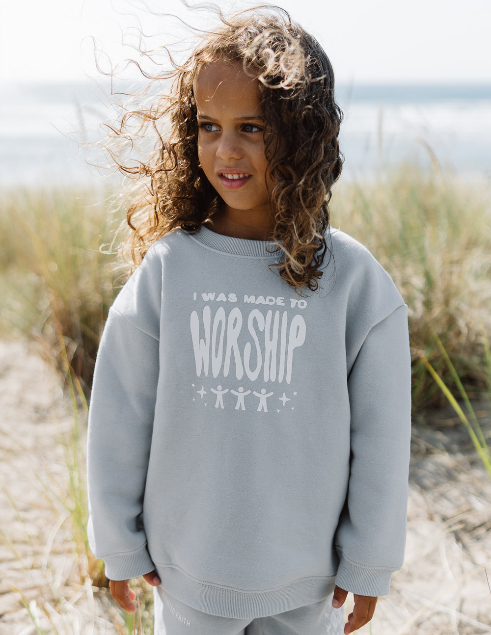 Child wearing a gray sweatshirt with 'I was made to worship' text on a beach.