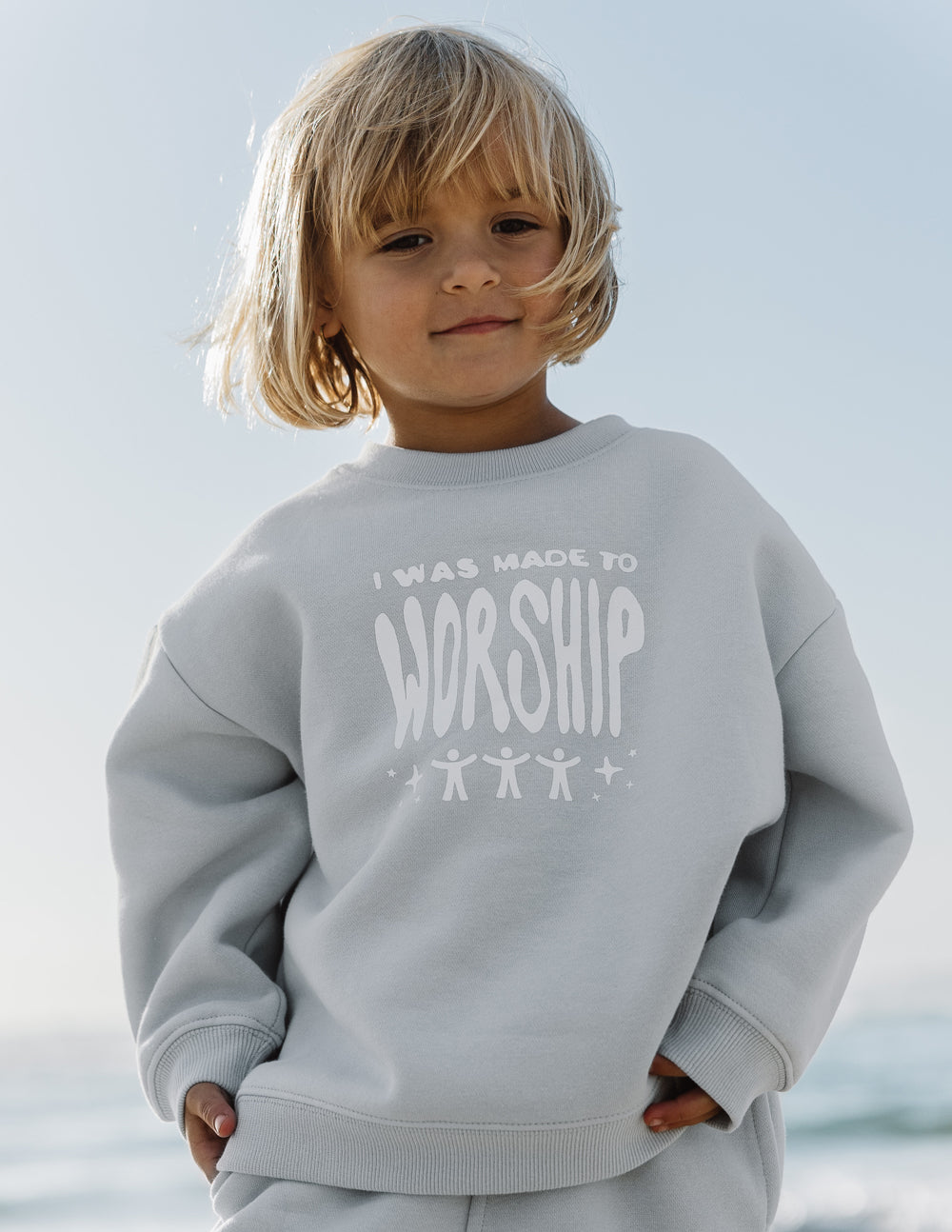Child wearing a gray sweatshirt with 'I was made to worship' text, standing against a light background.