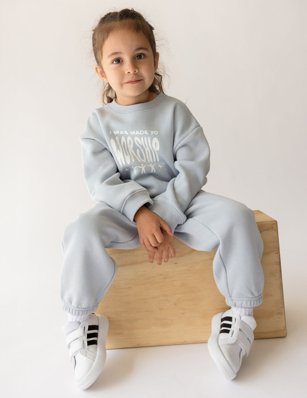 Child wearing a light gray sweatshirt and pants sitting on a wooden block against a white background
