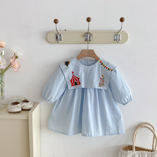 Light blue dress with animal and tent design on a wooden hanger against a white wall.