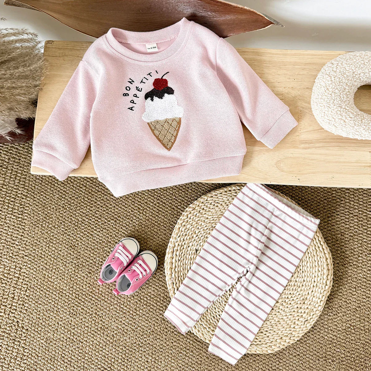 Pink children's sweatshirt with ice cream cone design, pink shoes, and striped pants on a wooden surface.