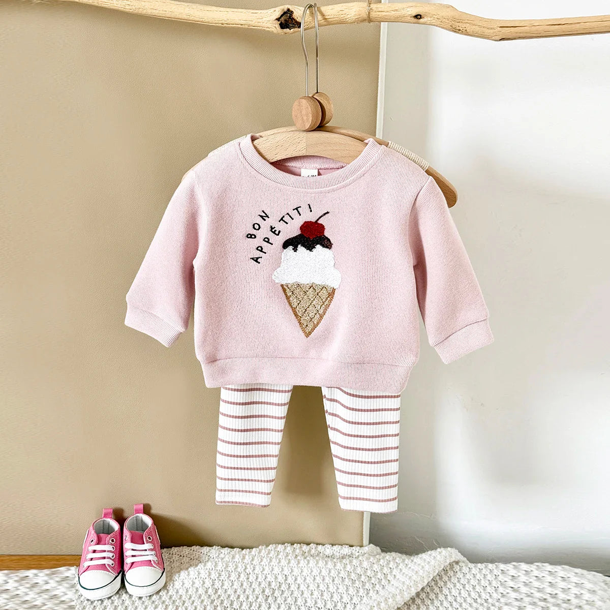 Pink children's outfit with ice cream design on a hanger against a beige wall.