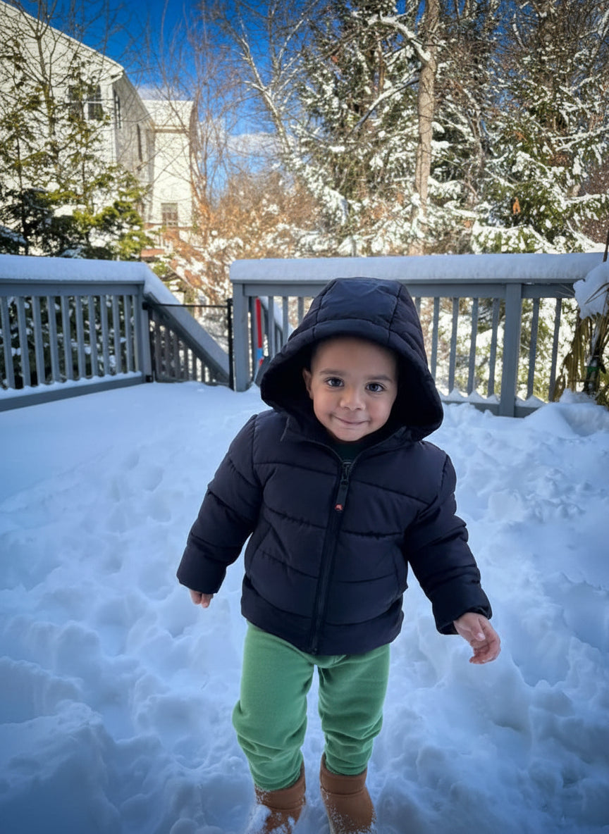 Child in a black coat and green pants standing on a snow-covered deck with trees in the background.