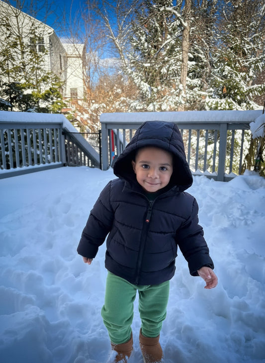 Child in a black coat and green pants standing on a snow-covered deck with trees in the background.