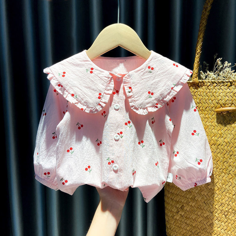Pink baby blouse with large ruffled collar and cherry print, held against dark curtain background