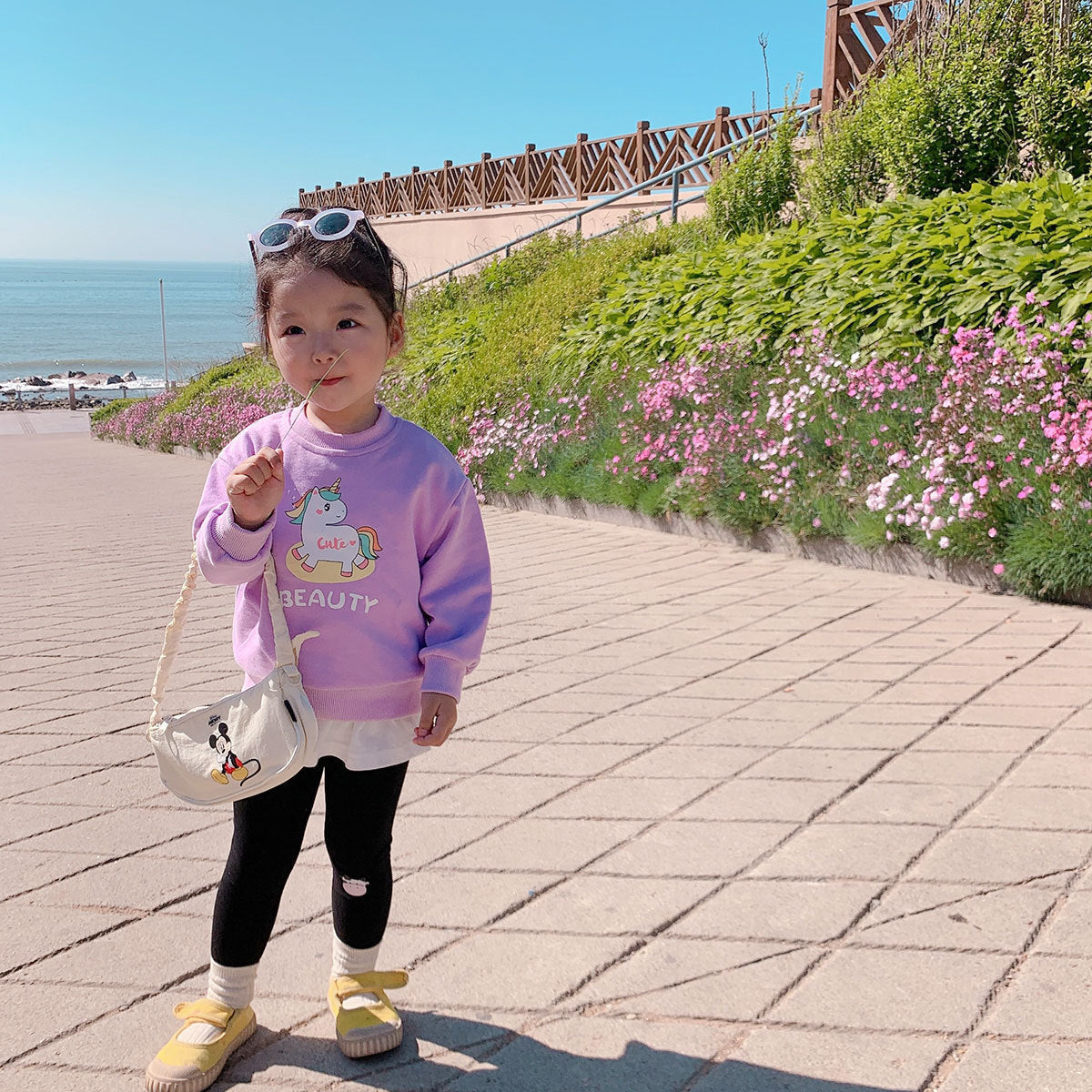 Little girl in pink unicorn sweater and black leggings standing by coastal path with flowers